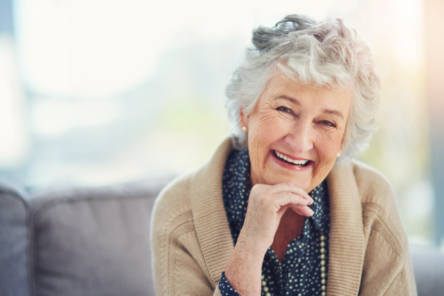 A smiling white haired elderly woman in a beige cardigan resting her chin on her hand.
