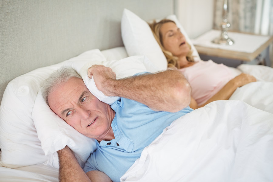 Man with a pillow over his ears to block out his wife's loud snoring due to sleep apnea.