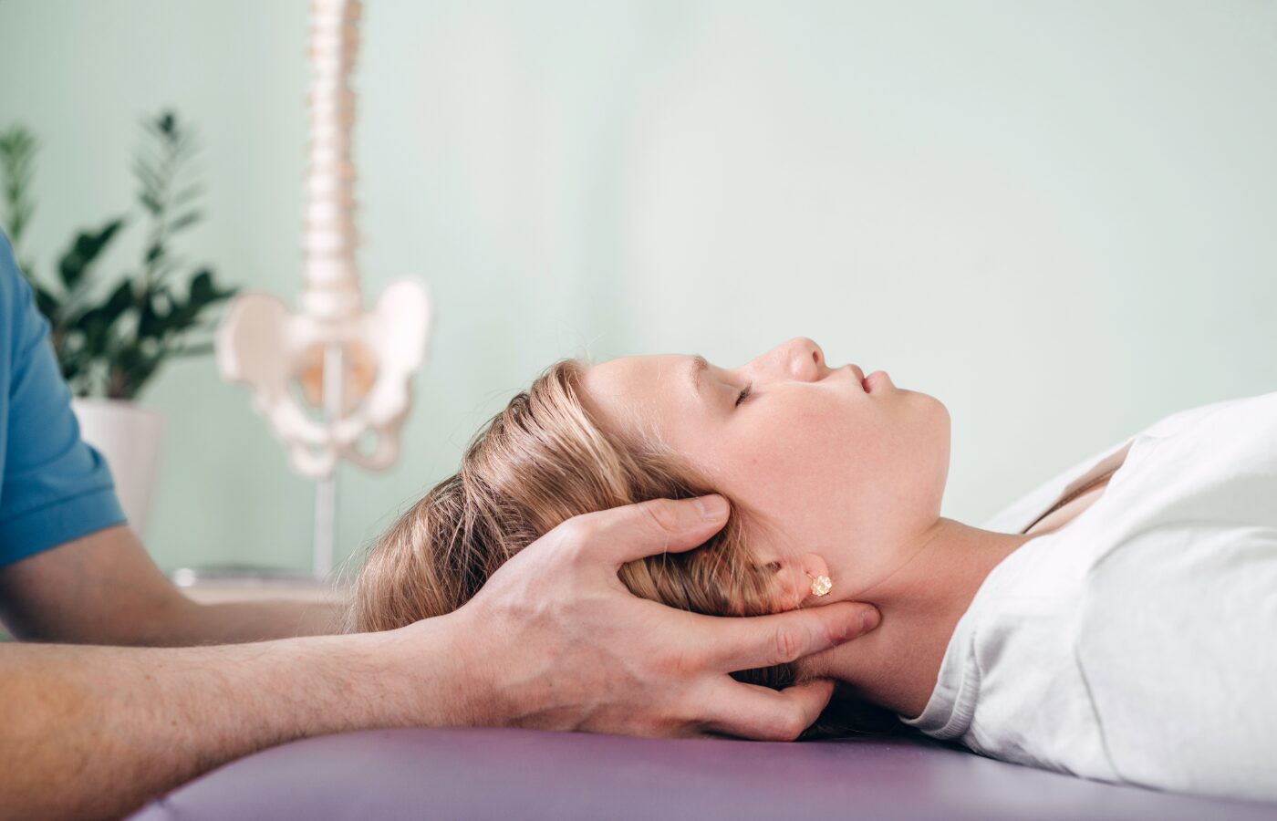 Therapist uses CraioSacral Therapy on a patient lying on the table with a model of the spine behind them.