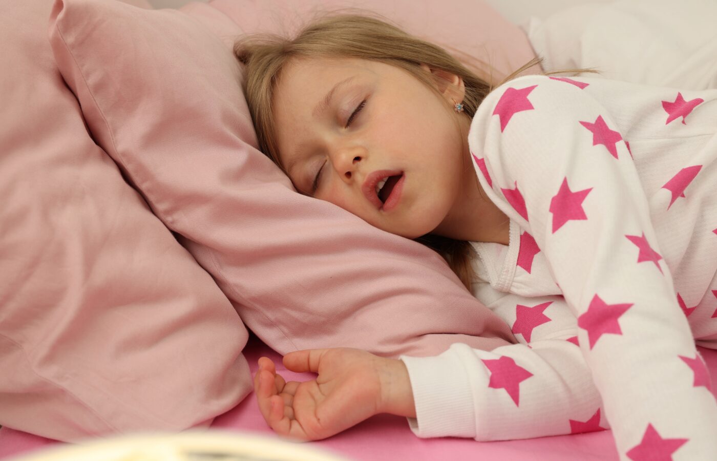 Photo of a young girl in white pjs with pink stars asleep on pink sheets with mouth open