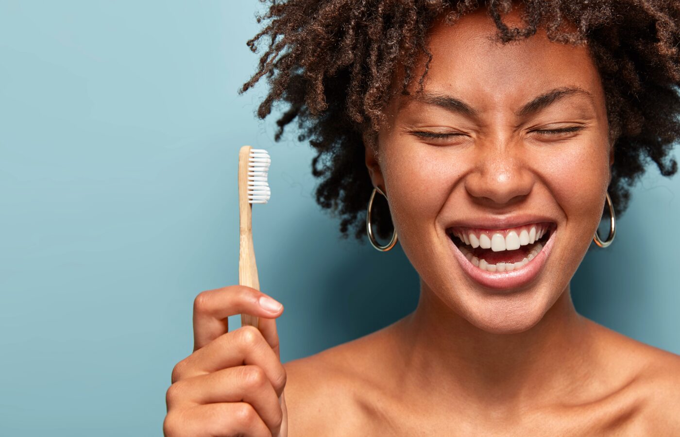 black woman against blue background holding a bamboo toothbrush with her eyes closed and a big smile with white teeth