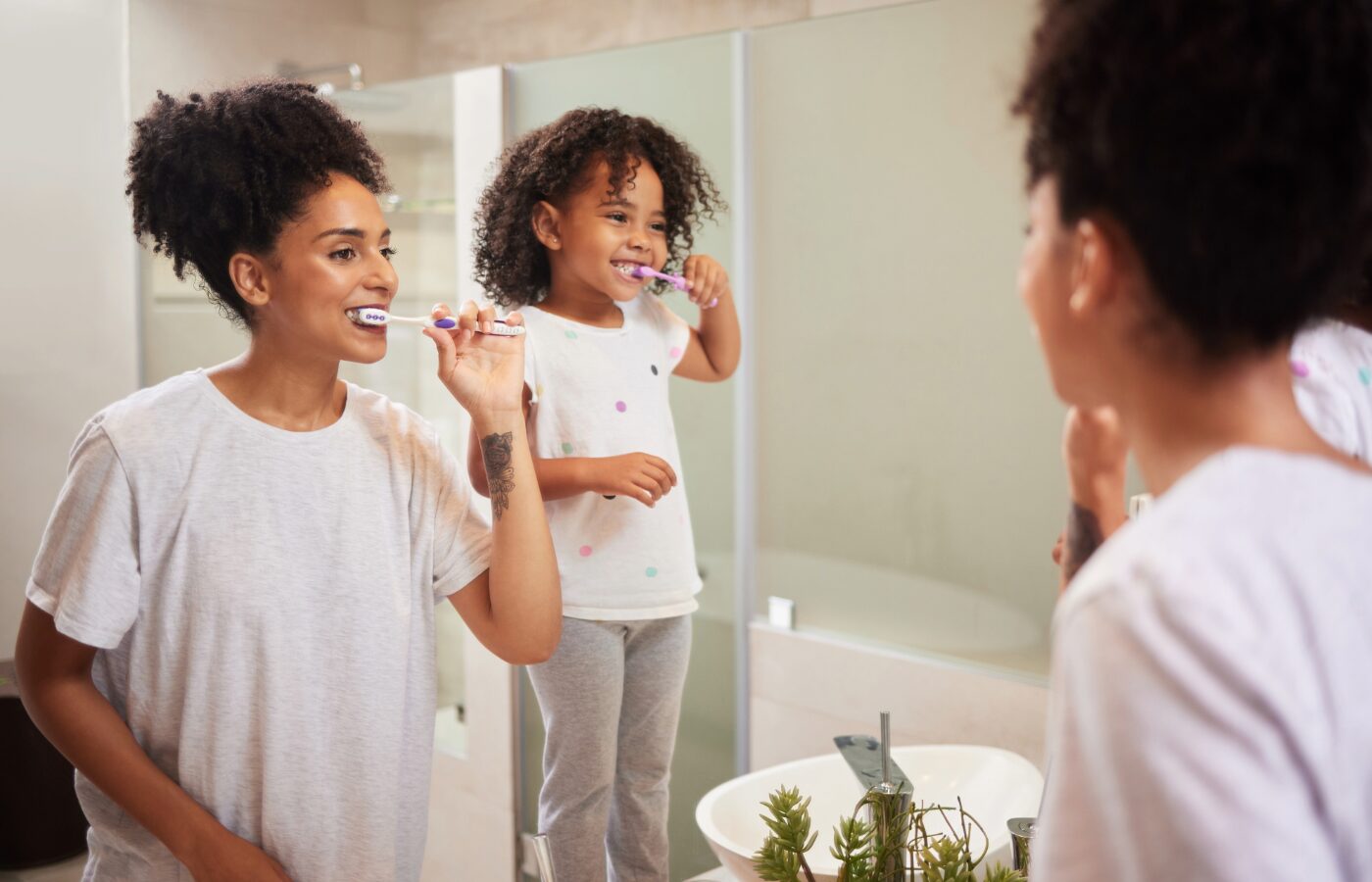 black mom and daughter standing in front of a mirror while brushing their teeth. You can see them in the right corner with their reflection in the mirror and toothbrushes in their mouths.