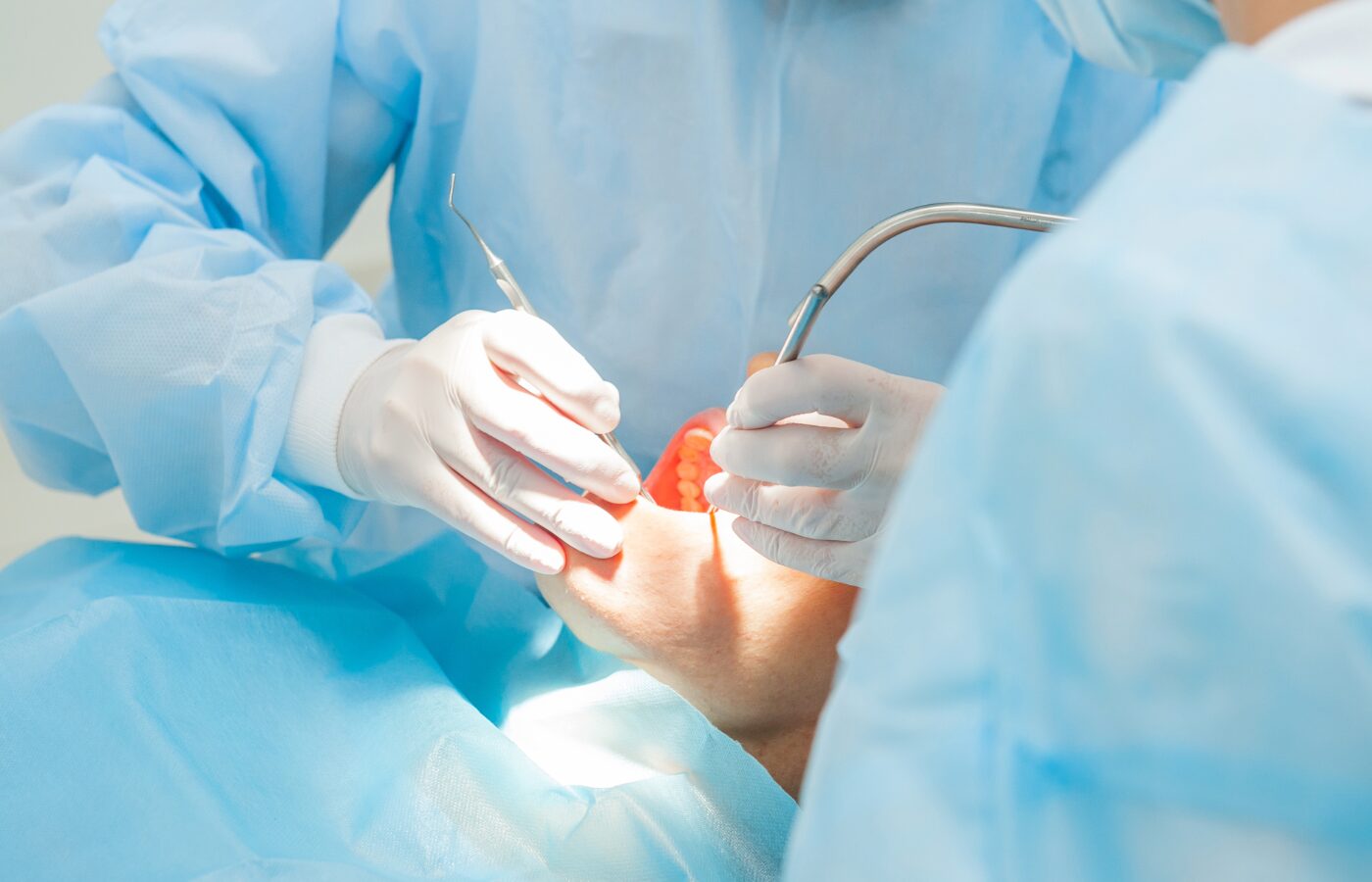 a dentist and dental assistant wearing dental gowns in blue surround a patient with his mouth open wide and instruments in his mouth.